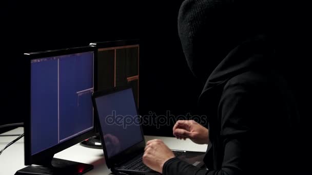 Хакер в Балаклаві зламує код за допомогою ноутбука та комп'ютерів зі своєї темної кімнати хакерів — стокове відео
