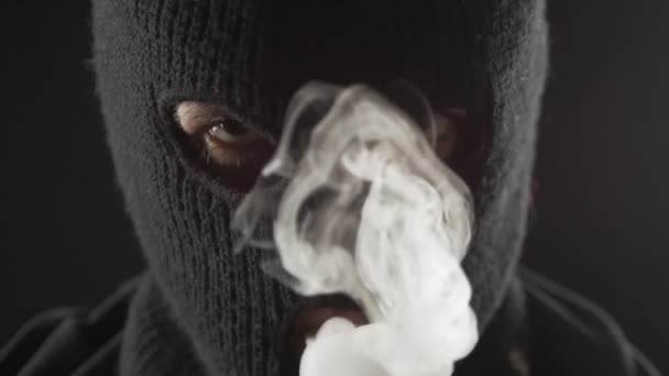 Terrorista peligroso en un pasamontañas fuma vapor de cigarrillo electrónico y hace mucho humo . — Vídeos de Stock