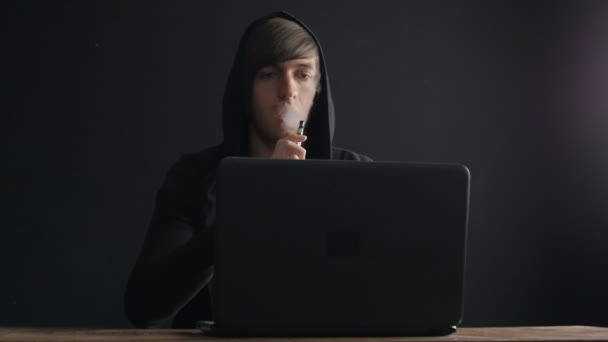 Человек-программист в капюшоне работает за ноутбуком и курит электронную сигарету — стоковое видео