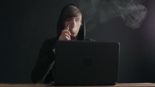 Man programmer in hood working at laptop and smoking e-cigarette — Stock Video
