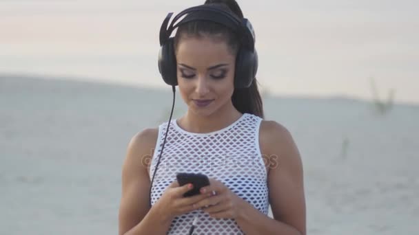 Mulher usando celular celular smartphone na praia usando fones de ouvido para música ou falando Menina de biquíni usando smartphone feliz . — Vídeo de Stock