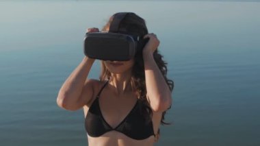 Bikinili genç kadın, sahilde bir sanal gerçeklik gözlük kullanır. Seksi kız yaz deniz arka plan üzerinde Vr-kulaklık kullanarak deneyim elde.