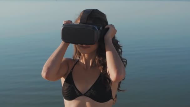 Jonge vrouw in bikini maakt gebruik van een virtual reality bril op het strand. Sexy meisje krijgt ervaring in het gebruik van Vr-headset op zomer op zee achtergrond. — Stockvideo