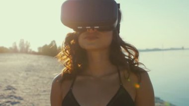 Bikinili genç kadın, sahilde bir sanal gerçeklik gözlük kullanır. Seksi kız yaz deniz arka plan üzerinde Vr-kulaklık kullanarak deneyim elde.