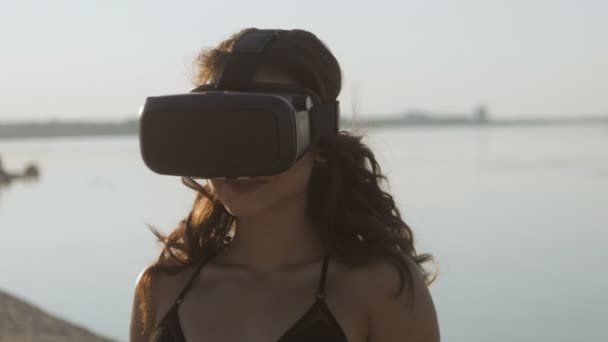 Bikinili genç kadın, sahilde bir sanal gerçeklik gözlük kullanır. Seksi kız yaz deniz arka plan üzerinde Vr-kulaklık kullanarak deneyim elde. — Stok video