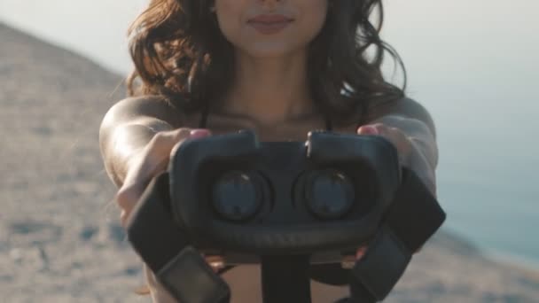 Młoda kobieta, dając zestaw wirtualnej rzeczywistości na plaży o zachodzie słońca. Dziewczyna na wakacjach daje okulary wirtualnej rzeczywistości z morzem — Wideo stockowe