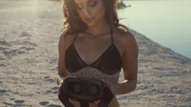 Jovem mulher sexy em biquíni dando fone de ouvido realidade virtual na praia ao pôr do sol. Menina em férias dá óculos de realidade virtual contra o mar — Vídeo de Stock