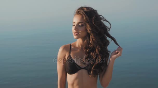 Mooie brunette meisje in een zwarte bikini op het strand op de achtergrond van de zee. — Stockvideo