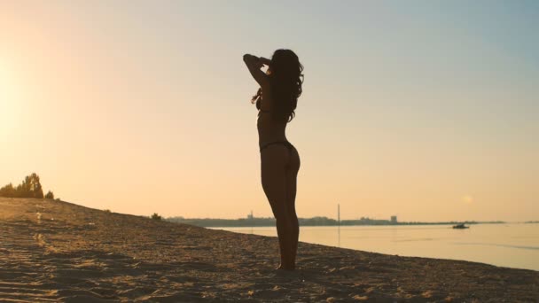 Ein Mädchen mit einem sexy Körper steht im Morgengrauen am Strand. junge glückliche Frau, die im Sand steht und sich gegen das Meer streckt. der Beginn eines guten Tages. — Stockvideo