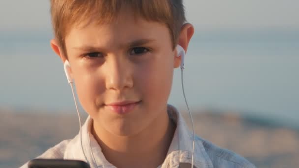 Rapaz de close-up a ouvir música nos auscultadores. Criança de 10 anos desfruta de um smartphone sentado na areia no fundo do mar — Vídeo de Stock