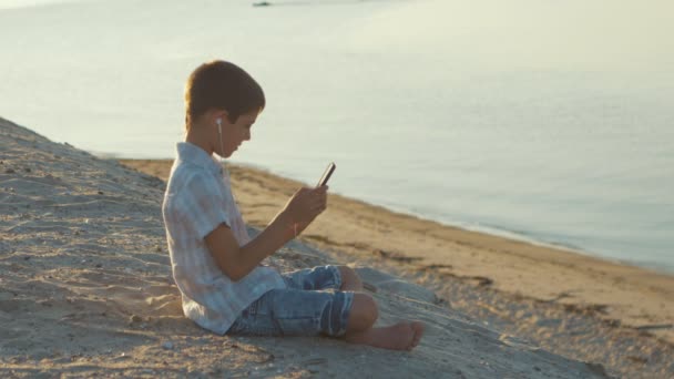 Retrato de un niño en la playa jugando con un smartphone. Niño de 10 años disfruta de un teléfono inteligente sentado en la arena en el fondo del mar — Vídeo de stock