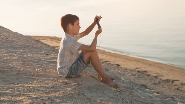 Portrett av en gutt på stranden som leker med en smarttelefon. Ti år gamle barn nyter en smarttelefon sittende på sanden i bakgrunnen av havet – stockvideo