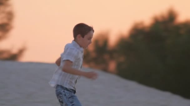 Χαριτωμένο αγόρι τρέχει και πηδά από ένα λόφο άμμο στο ηλιοβασίλεμα. Ενεργό υπόλοιπο του παιδιού. — Αρχείο Βίντεο