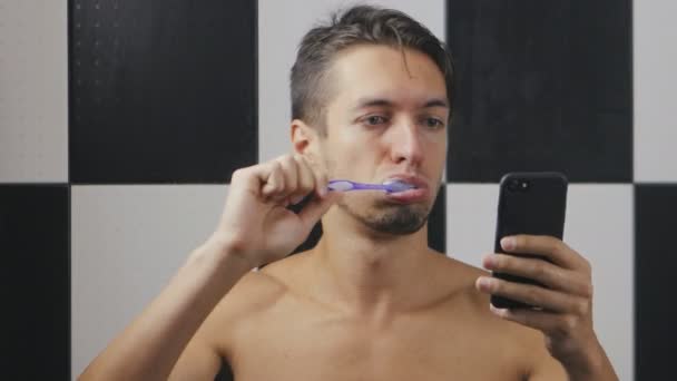 Занятой бизнесмен в ванной чистит зубы и пользуется смартфоном — стоковое видео