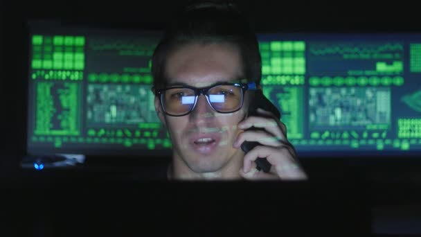 Хакер-программист в очках разговаривает по телефону в центре кибербезопасности, заполненном дисплейными экранами . — стоковое видео
