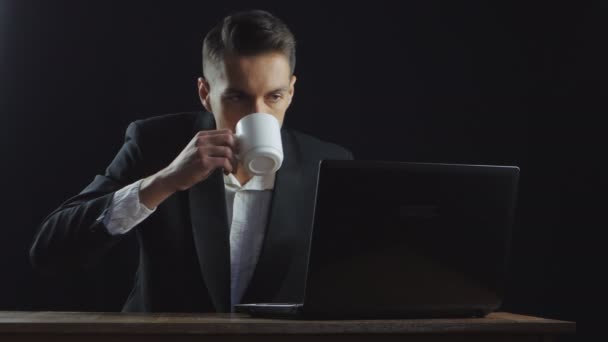 青年実業家は、暗いオフィスの部屋でラップトップで働きながらコーヒーを飲む — ストック動画