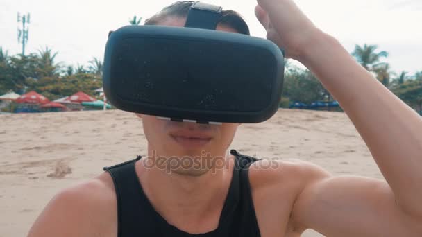 Молодой человек пользуется очками виртуальной реальности на пляже. Парень получает опыт использования гарнитуры во время летних путешествий — стоковое видео
