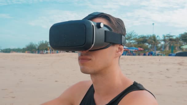 Молодой человек пользуется очками виртуальной реальности на пляже. Парень получает опыт использования гарнитуры во время летних путешествий — стоковое видео