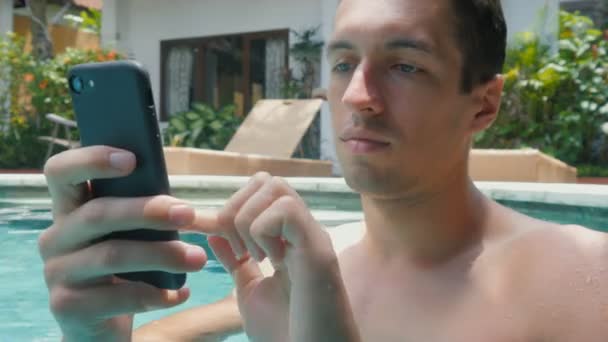 Jovem usa um smartphone enquanto está na piscina do resort — Vídeo de Stock