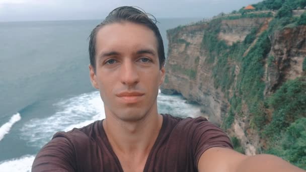 Man maakt selfie tegen de achtergrond van een rotsachtige kust en de oceaan golven — Stockvideo