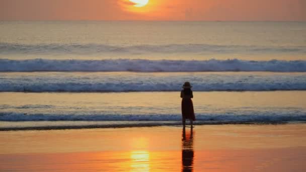 Silhouette de femme en chapeau à la plage au bord de l'océan. Femme debout dans les vagues et profitant du magnifique coucher de soleil 
