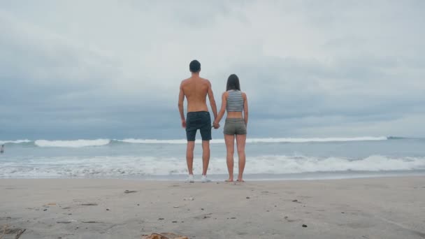 Joven familia cogida de la mano se para frente al océano en la playa. Feliz pareja joven disfrutando de las olas del océano durante las vacaciones de viaje — Vídeos de Stock