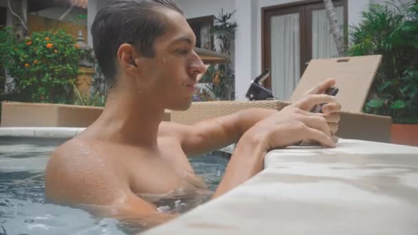 Hombre joven nada en la piscina y charlando en el teléfono inteligente en las vacaciones de verano — Vídeos de Stock