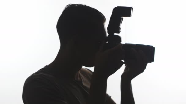 Фотограф Силуэт крупный план. Молодой человек фотографирует на белом фоне с помощью камеры DSLR — стоковое видео
