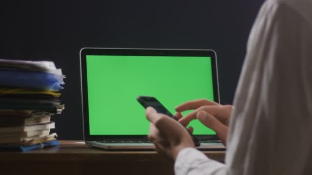 人类使用的是智能手机, 他的笔记本电脑上有一个绿色的屏幕, 或者 chromakey 在他的桌面上。生意人晚上在办公室工作. — 图库视频影像