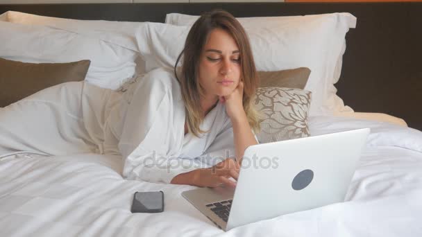 Junge Frau im Bademantel auf Bett liegend und am Laptop arbeitend — Stockvideo