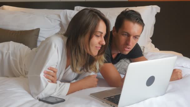 Νεαρό ζευγάρι στο μπουρνούζι ξαπλωμένος στο κρεβάτι και να εργάζονται στο lap-top — Αρχείο Βίντεο