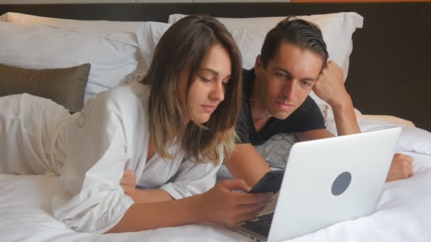 Young couple in bathrobe lying on bed and working on laptop — Stock Video