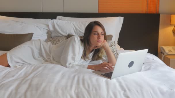 Young woman in bathrobe lying on bed and working on laptop — Stock Video