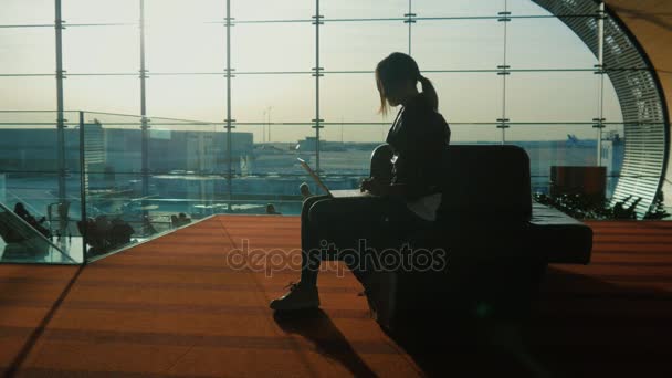 Молода жінка працює з ноутбуком в терміналі аеропорту. Чекаю на мій рейс. Силует на тлі великого вікна — стокове відео