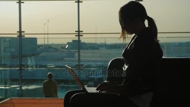 Mujer joven que trabaja con el ordenador portátil en la terminal del aeropuerto. Esperando mi vuelo. Silueta sobre el fondo de una gran ventana — Vídeo de stock