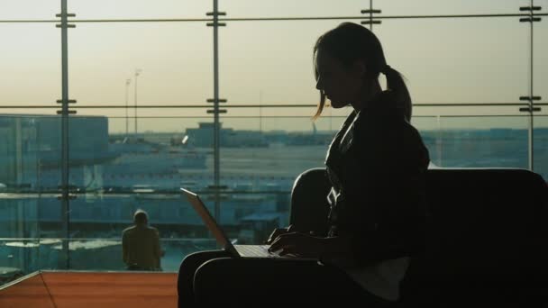 Mujer joven que trabaja con el ordenador portátil en la terminal del aeropuerto. Esperando mi vuelo. Silueta sobre el fondo de una gran ventana — Vídeo de stock