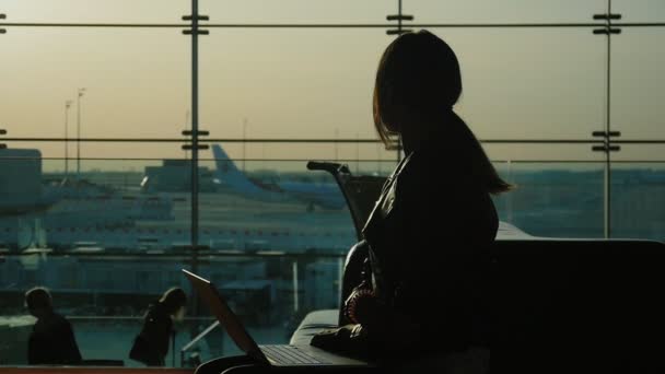 Young woman working with laptop in airport terminal. Waiting for my flight. Silhouette against the background of a large window — Wideo stockowe