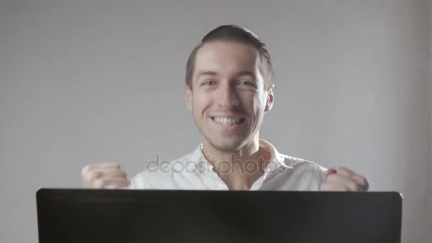 Young man in a white shirt very happy and rejoices sitting at laptop in office — Stock Video