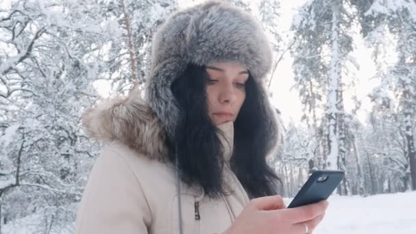 Närbild av ung kaukasisk kvinna i en varm mössa med en smartphone i vinter snöig skog — Stockvideo