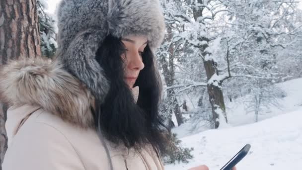Primer plano de la joven mujer caucásica en un sombrero caliente usando un teléfono inteligente en el bosque nevado de invierno — Vídeo de stock