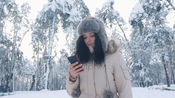 Young Caucasian Woman in a warm hat using a smartphone in winter snowy forest — Stock Video