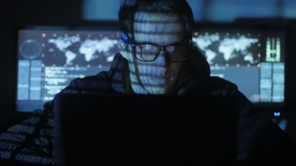 Programador Hacker en gafas está trabajando en la computadora, mientras que los caracteres de código azul reflejan en su cara en el centro de seguridad cibernética lleno de pantallas de visualización . — Vídeos de Stock