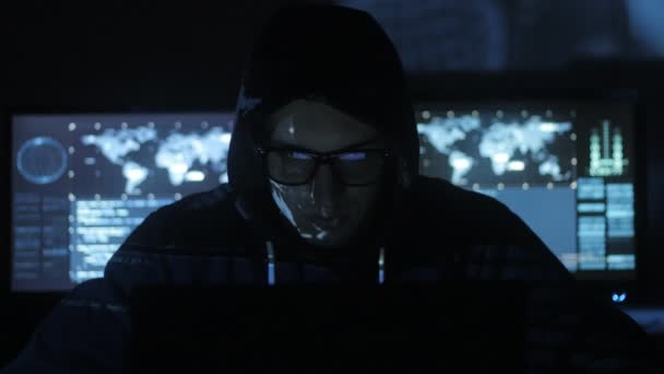 Man geek hacker in hood with glasses working at computer while blue code characters reflect on his face in cyber security center filled with display screens. — Stock Video