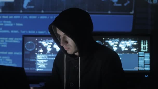 Mannen geek hacker i hood arbetar vid dator medan blå kod tecken reflektera över hans ansikte i cyber security center fylld med skärmar. — Stockvideo