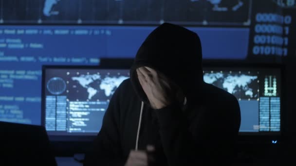 Hombre geek hacker en el capó trabajando en exceso en la computadora y sufre de un dolor de cabeza en el centro de seguridad cibernética lleno de pantallas de visualización . — Vídeos de Stock