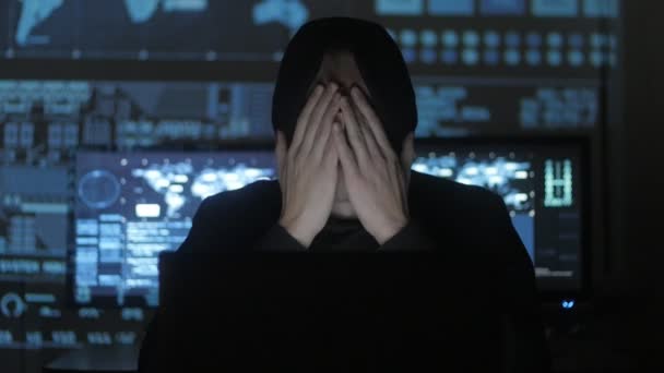Man geek hacker in hood overworking at computer and suffers from a headache in cyber security center filled with display screens. — Stock Video
