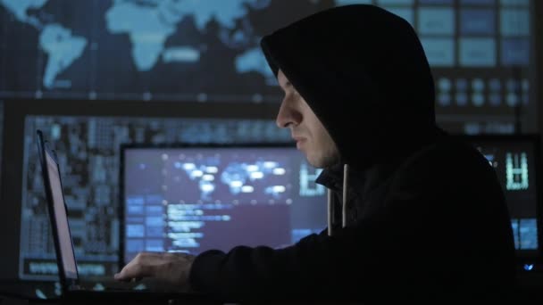 Mannen geek hacker i hood arbetar på dator i cyber security center fylld med skärmar. — Stockvideo