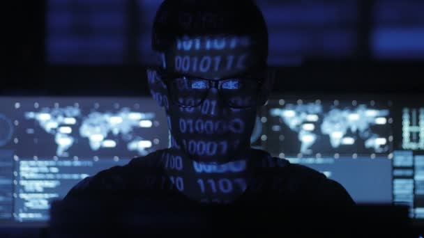 Programmeur pirate dans les lunettes travaille sur ordinateur tandis que les caractères de code binaire bleu réfléchissent sur son visage dans le centre de cybersécurité rempli d'écrans d'affichage .