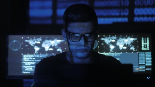 Hacker programmatore in occhiali sta lavorando sul computer, mentre blu caratteri di codice binario riflettere sul suo volto nel centro di sicurezza informatica pieno di schermi di visualizzazione . — Video Stock