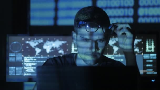 Hacker programmer in glasses is working on computer while blue binary code characters reflect on his face in cyber security center filled with display screens. — Stock Video